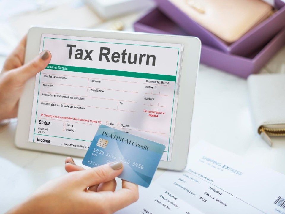 Tablet screen shows a tax return form while a blue credit card is held in the foreground, ready for payment.